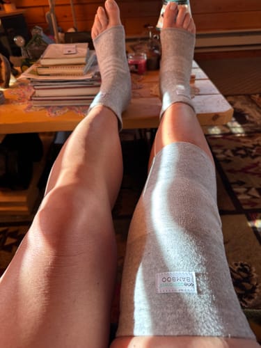 Customer resting their legs while wearing the grey bamboo footsleeves and a matching knee sleeve.