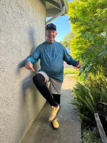 image 3 out of 3: Customer smiling outdoors while pointing to the black compression sock on their raised leg.
