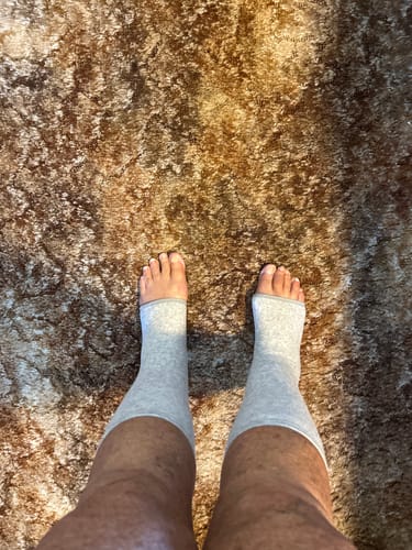 Reviewer standing while wearing the light grey, open-toed anti-fatigue footsleeves on their feet.