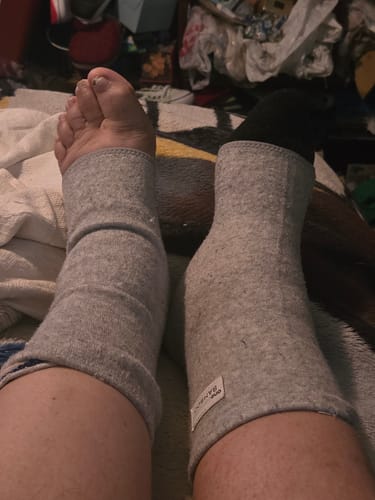 Reviewer wearing the gray Anti-Fatigue Bamboo Footsleeves on both legs, one over a sock, while resting.