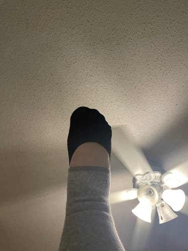 A customer's foot pointing upwards, wearing the black anti-fatigue footsleeve over a grey sock indoors.